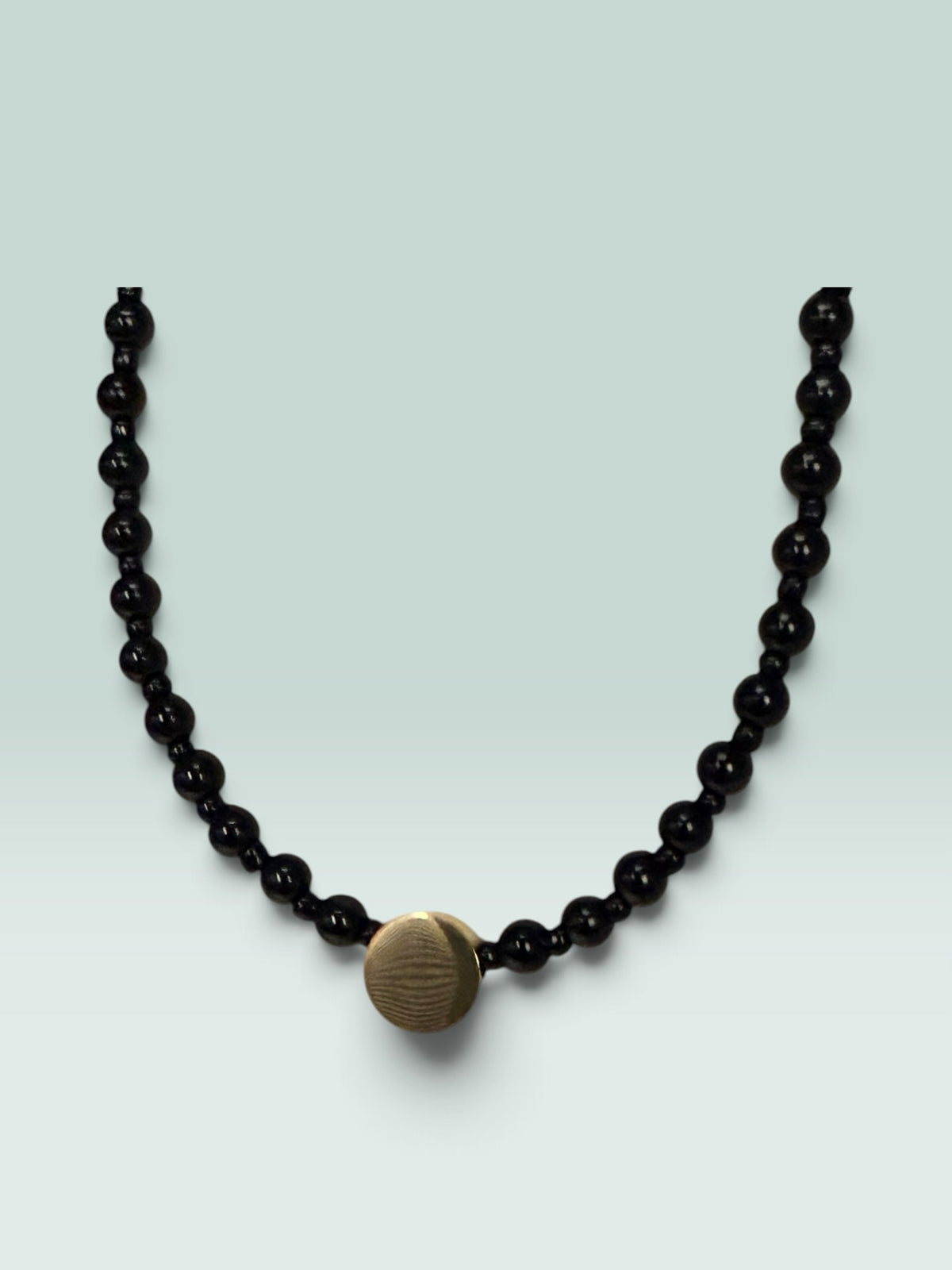 The Salty Bead Co minimalist necklace featuring polished black beads and a textured gold-tone center disc accent.