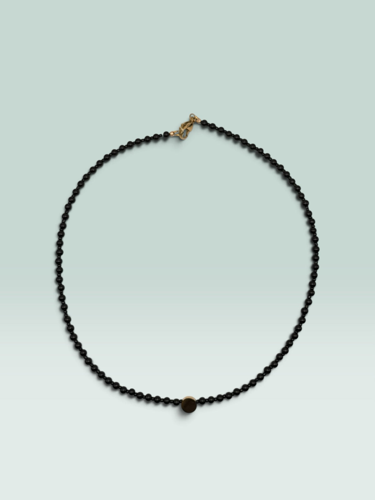 The Salty Bead Co minimalist black beaded necklace featuring a small textured gold-tone center disc accent