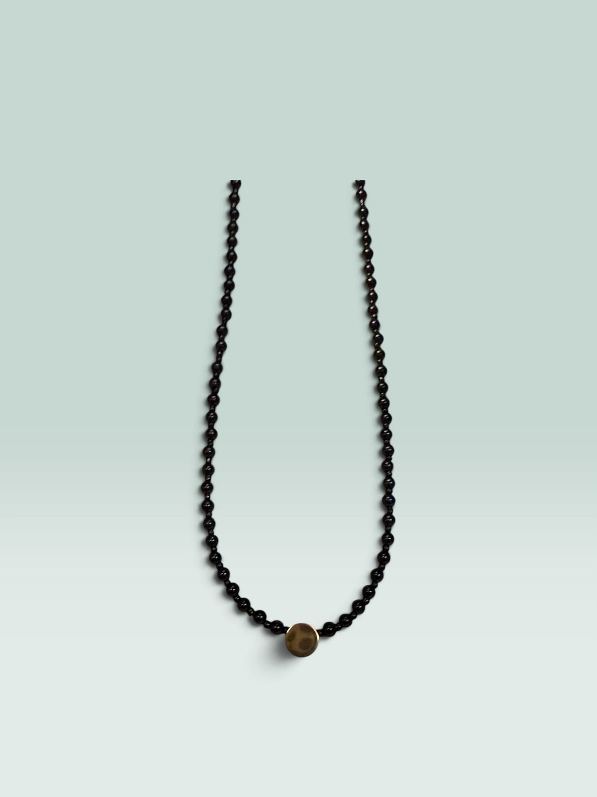 The Salty Bead Co minimalist necklace featuring polished black beads and a central textured gold-tone disc accent.