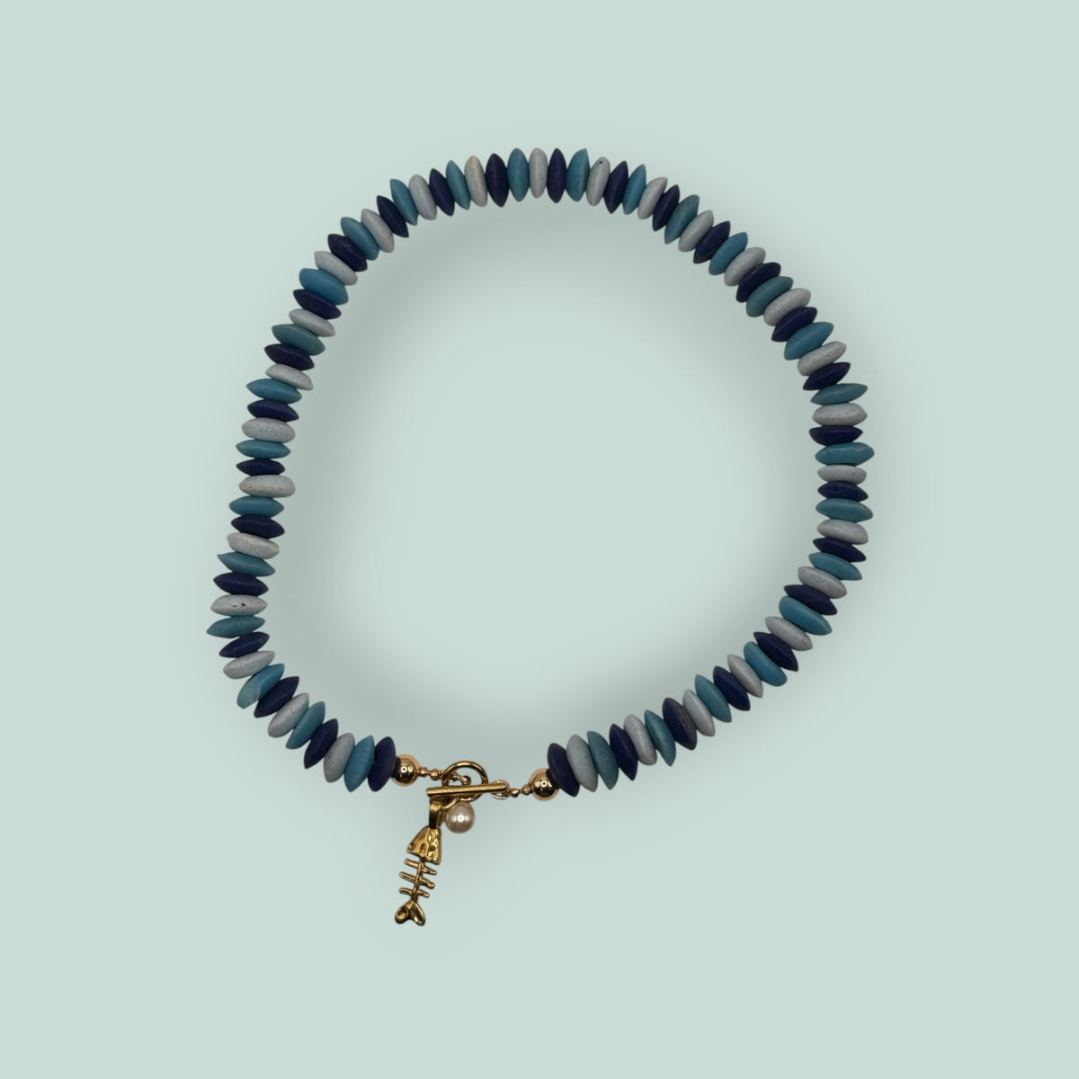 The Salty Bead Co handcrafted necklace featuring blue disc beads, a gold fish skeleton charm, and a faux pearl accent.