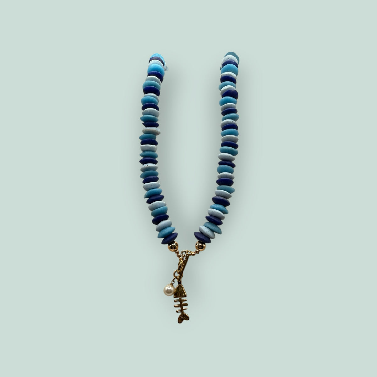 The Salty Bead Co handcrafted necklace with blue disc beads, gold toggle clasp, fish skeleton charm, and faux pearl.