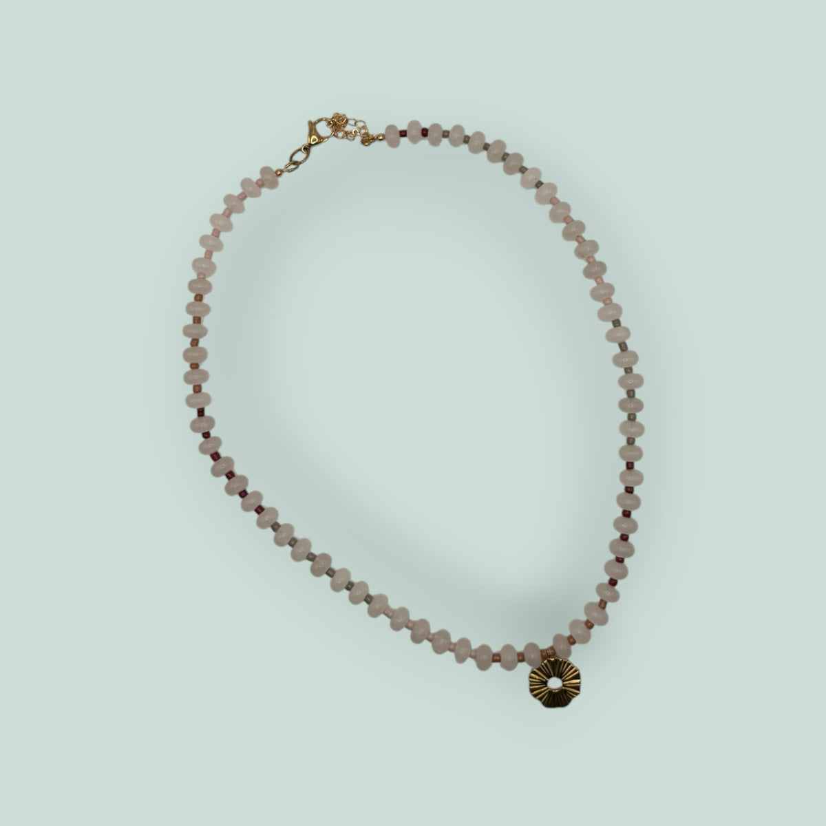 The Salty Bead Co handcrafted necklace with pale pink gemstone beads, dark red accents, and a gold sunburst pendant.