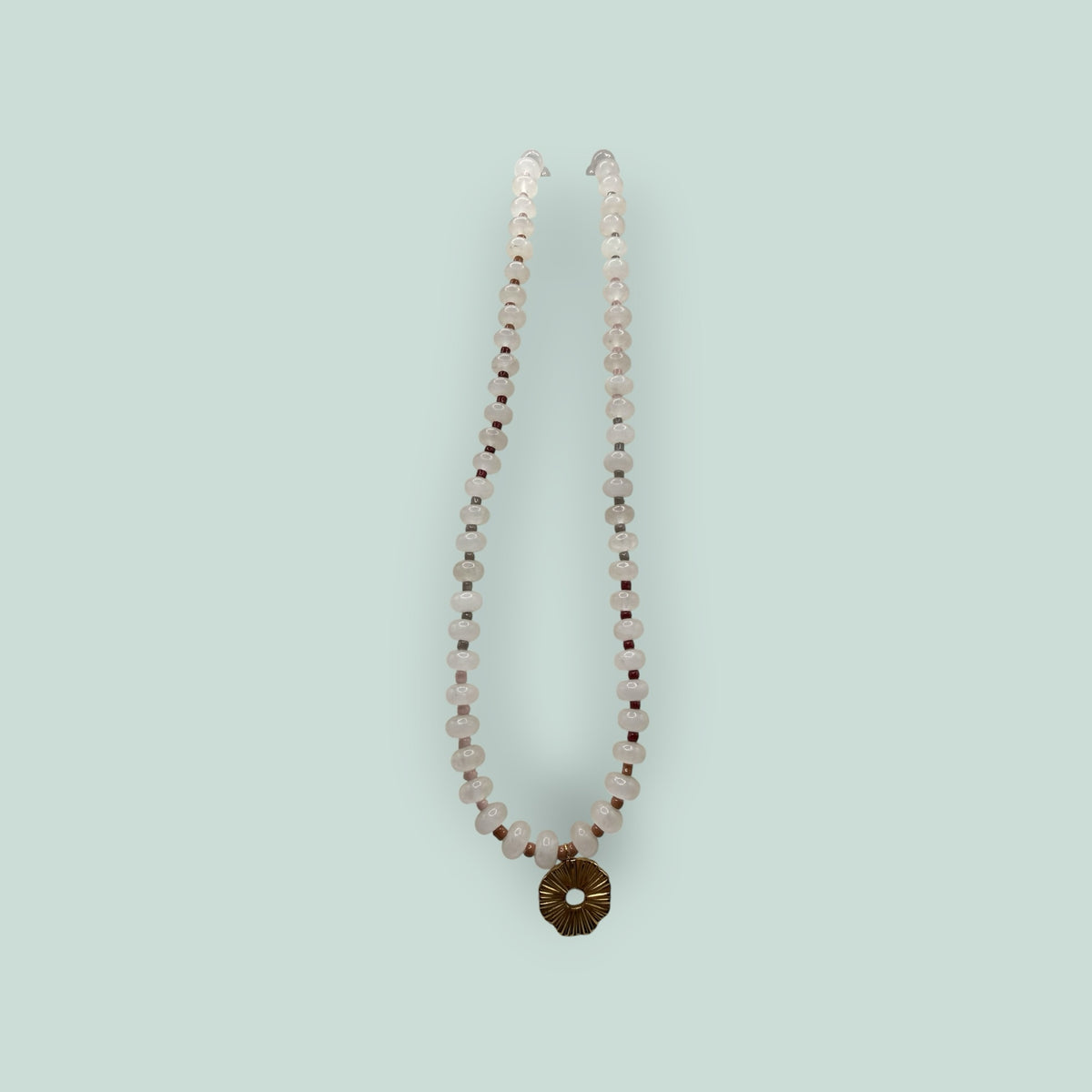 The Salty Bead Co handcrafted necklace with pale pink gemstone beads, dark red accents, and a gold sunburst pendant.