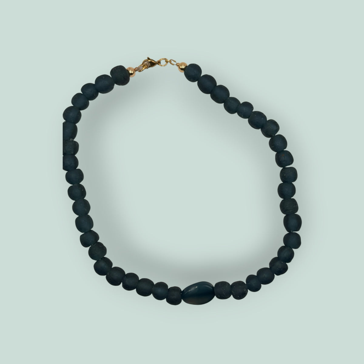 The Salty Bead Co handcrafted necklace with matte teal-blue beads and a polished oval stone centerpiece, gold clasp.