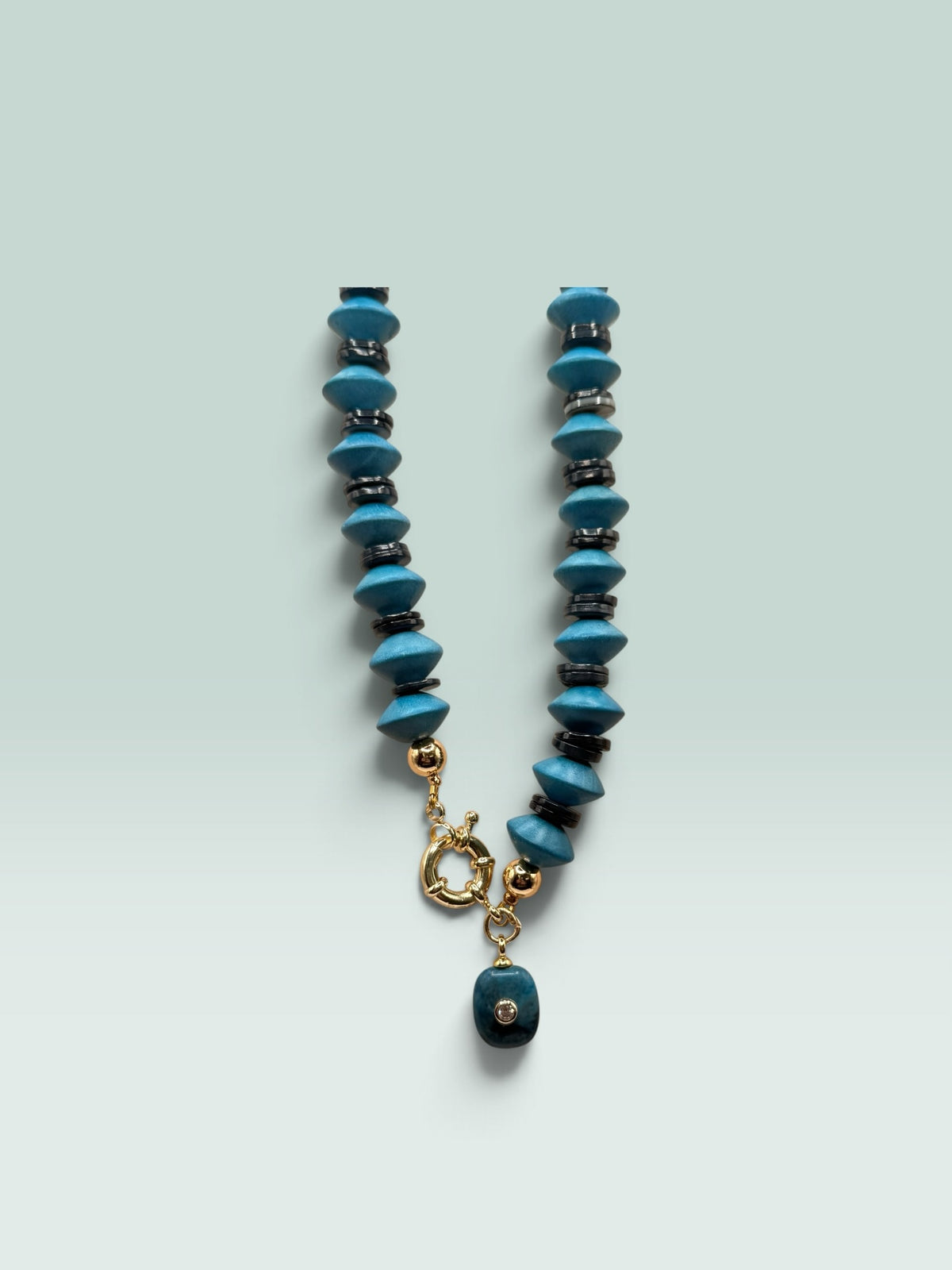 The Salty Bead Co handcrafted statement necklace with turquoise-blue disc beads, dark metallic spacers, and gold clasp.